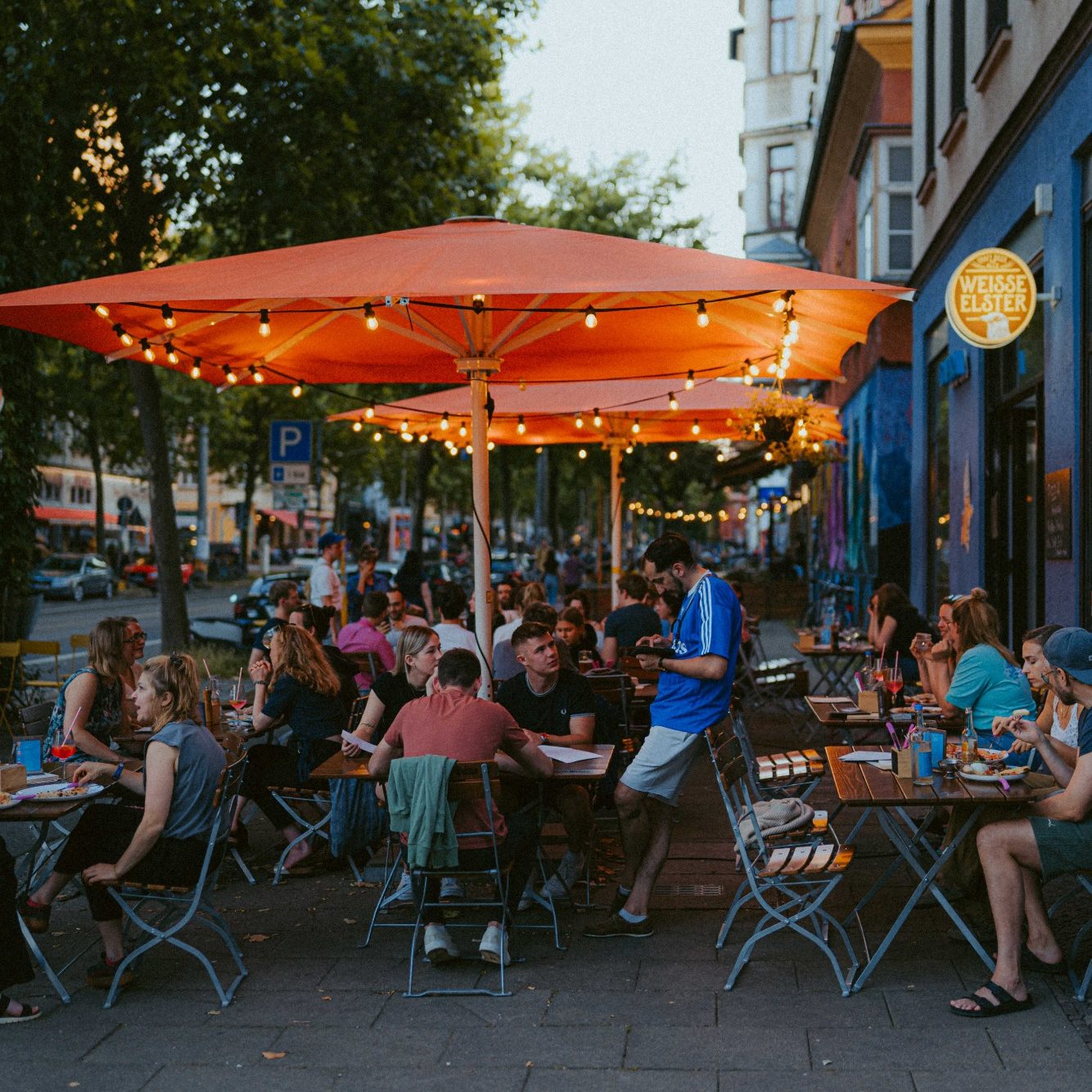 Ein früher Sommerabend auf der Terrasse des Capra. Abendsonne, grüne Bäume, orangene Sonnenschirme mit Lichterketten, der Außensitz ist voll