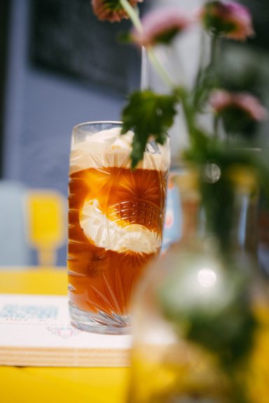 Ein Glas Eiskaffee hinter einer Blumenvase auf einem gelben Tisch. Sommer-Feeling.