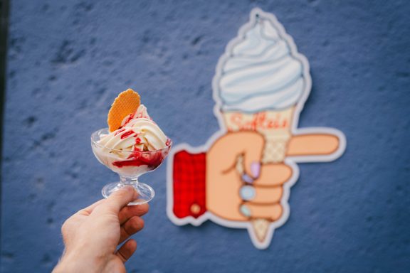 Eine Schale Eis mit Erdbeersoße und Waffel wird für ein Schild gehalten, das eine Eistüte zeigt, auf der "Softeis" steht.